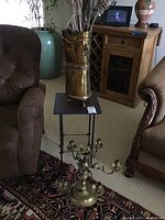 Full view of the copper vase with twigs and the brass candelabra positioned on the floor near chairs and a small table.