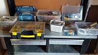 Overview of multiple plastic toolboxes and containers filled with screws, nails, wall anchors, wire, and various hardware components on a metal table.