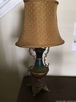 Full side view of one vintage table lamp showing beige shade, decorative vase body with green and brown gradient, heavy ornate metal base with claw feet.