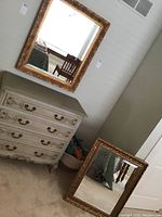 Two mirrors shown, one hanging on the wall above a three-drawer chest and another resting on the floor. Both feature elaborate gold-colored frames with vine and leaf designs.