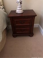 Wooden nightstand with two drawers and metal handles next to a bed, showing front and side views.
