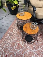 Two round side tables with wooden tops and black metal wire bases, placed on a patterned outdoor rug next to patio furniture. Displayed with two glass solar lantern lights.