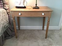 Front view of distressed wooden table with turned legs and single drawer with white knobs.