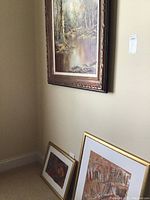 Framed landscape painting hung on wall above two other framed artworks leaning against baseboard