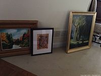 Photo showing four framed artworks on the floor: two larger paintings on canvas and two smaller prints.