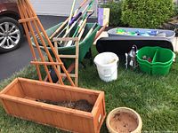 Green wheelbarrow with gardening tools including rake and shovels arranged inside, wooden rectangular planter box with trellis filled with soil, round terracotta planter pot, white bucket, green gardening caddy, and grey plastic tote seen on grass in front of house.