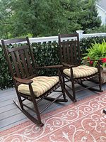 Two dark brown wooden rocking chairs on a deck, each with a striped cushioned seat and visible scratch marks on wood.
