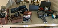 Photo showing multiple vintage cameras and camera cases arranged on a table, including Kodak, Polaroid, and Bell and Howell cameras, with cases and flash unit.