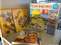 Vintage Kenner Easy-Bake Oven in original box, two yellow metal turtle crossing signs, Jim Davis Garfield quartz wall clock displayed together.