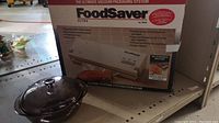 Foodsaver Ultra vacuum packaging system in original box with Pyrex brown glass lidded casserole placed beside it on a shelf.