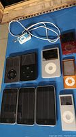 Photo showing 12 digital music players on a blue surface including iPod Classics, iPod Nanos, and iPod Touch devices in various colors.
