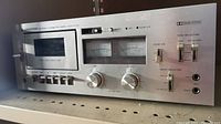 Front angle view showing the cassette compartment, analog VU meters, and control buttons on the silver front panel of the Kenwood stereo cassette deck KX-830