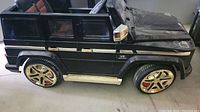 Side view of the black kids electric Mercedes G55 AMG toy vehicle showing two doors, gold accents on wheels and side steps, and brown-black seats.