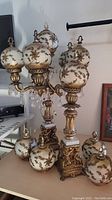 Photo showing two ornate metal and marble base lamps with white glass globes decorated with raised gold-tone leaf and flower designs. One lamp has five globes with hanging glass lusters, the other has a single globe.