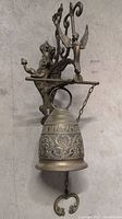 Side view of brass monastery wall bell mounted on ornate bracket with angel figures and scrollwork, pull chain hanging below