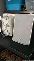 Photo of two outdoor speakers showing front and back sides, mounting hardware visible on back of one speaker.