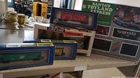 Photo showing multiple boxed toy train cars including Chessie green boxcar, Penn Central yellow gondola, and Life-Line evergreen trees with Santa's Toyland Express Deluxe train set box in background.