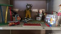 Shelf photo showing all vintage toys in lot including the Tonka crane, bear plush on skates, Piggy Cook, Peter Pig pull toy, and tin watercolors case.