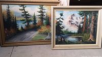 Two framed paintings leaning against a wall on the floor, showing both artworks together.