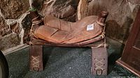 Side view of antique saddle stool showing leather seat and rawhide wrapped wood frame with rope lacing.