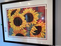 Framed art print showing vibrant sunflowers with three large blossoms against red and green leaves background.