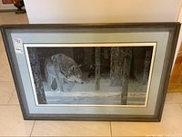 Full view of framed print showing wolf walking through snow with forest background, in wooden frame with gray mat.