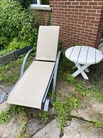 Chaise lounge with adjustable back and metal frame, beige mesh fabric seat, placed outdoors with plants and brick wall background, next to a round white painted wood table.