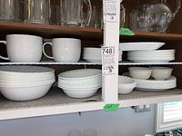 Wide view of white Corelle Vitrelle dinnerware set showing mugs on top shelf, bowls, salad plates and dinner plates stacked underneath on a shelf with floral liner.