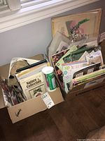 Two cardboard boxes filled with various needlecraft and craft instruction materials seen from above, showing assorted booklets, kits, and papers.