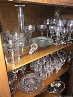 Full view of two wooden shelves containing etched glass stemware including wine glasses, coupes, a decanter with stopper, drinking glasses, and glass plates.