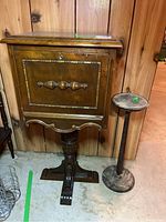 Front view of wood humidor cabinet on carved pedestal stand, and smaller round pedestal stand next to it.