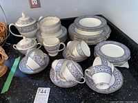 Full view of the china set showing stacked plates, bowls, tea cups, saucers, creamer, sugar bowl, and teapot displayed on black countertop.