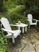 Two white Muskoka chairs positioned on a stone patio with a white square wooden side table between them, green foliage background. Stone sculpture on table not included.