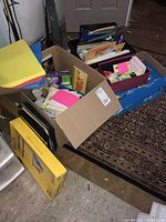Wide view of three boxes filled with assorted office supplies including paper stacks, sticky notes, folders, and packaging boxes.