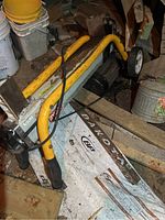 Electric log splitter showing yellow tubular protective handle bars, Ryobi brand label, electrical cord, and wheels at one end. Rust and dirt visible on metal surfaces.