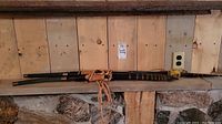 Wide view of both swords side by side on wood shelf against plank wall and stone background