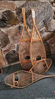 Two pairs of vintage-style wooden snow shoes with rawhide weaving, one larger pair standing upright against a rocky stone background and one smaller pair laid flat.