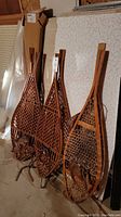 Three pairs of vintage wooden snow shoes leaning against a wall, showing the lattice woodwork and leather straps.