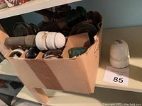 Box containing around a dozen ceramic electrical insulators in white, black, and green, plus one large white insulator placed next to the box.