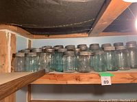 Top shelf of wooden shelving holding 13 antique Mason jars with metal lids, glass in clear and blue-green tones.