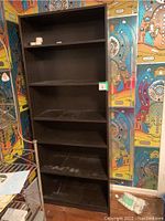 Front view of one black bookshelf with five shelves, showing some light wear and dust, against colorful wall.