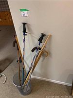 Overall view of seven canes and walking sticks placed inside a cylindrical holder next to a wall in an indoor setting, showing the variety of handle styles and materials.