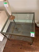 Square glass top table with metal frame and lower glass shelf next to a sofa, showing size and condition.