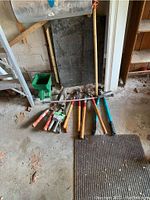 Assorted garden hand tools laid out on the floor and leaning against a wall, including trowels, pruners, and digging tools.