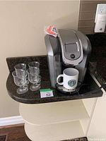 Full view of the Keurig 2.0 coffee maker with white Starbucks mug placed on drip tray, four glass coffee cups displayed alongside on a countertop.