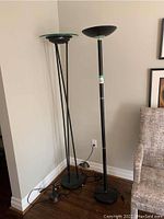 Photo of two standing torchiere lamps placed side-by-side against the wall, showing tall black metal bases and bowl-shaped shades with one having a green glass accent.