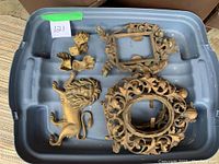 Top-down photo of a plastic storage bin containing various metal decorative pieces including two ornate metal frames, a lion wall accent, several small hand-shaped decorations, and other metal accents. Items show wear and vintage patina.
