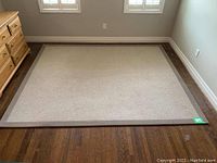 Full view of rectangular beige rug with brown border on hardwood floor near dresser.