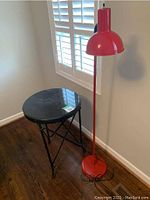 Black round side table and red floor lamp against a white wall with plantation shutters in the background.