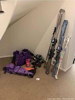 Full lot showing skis, poles, bags, boots and accessories on floor and leaning on wall under stairs.
